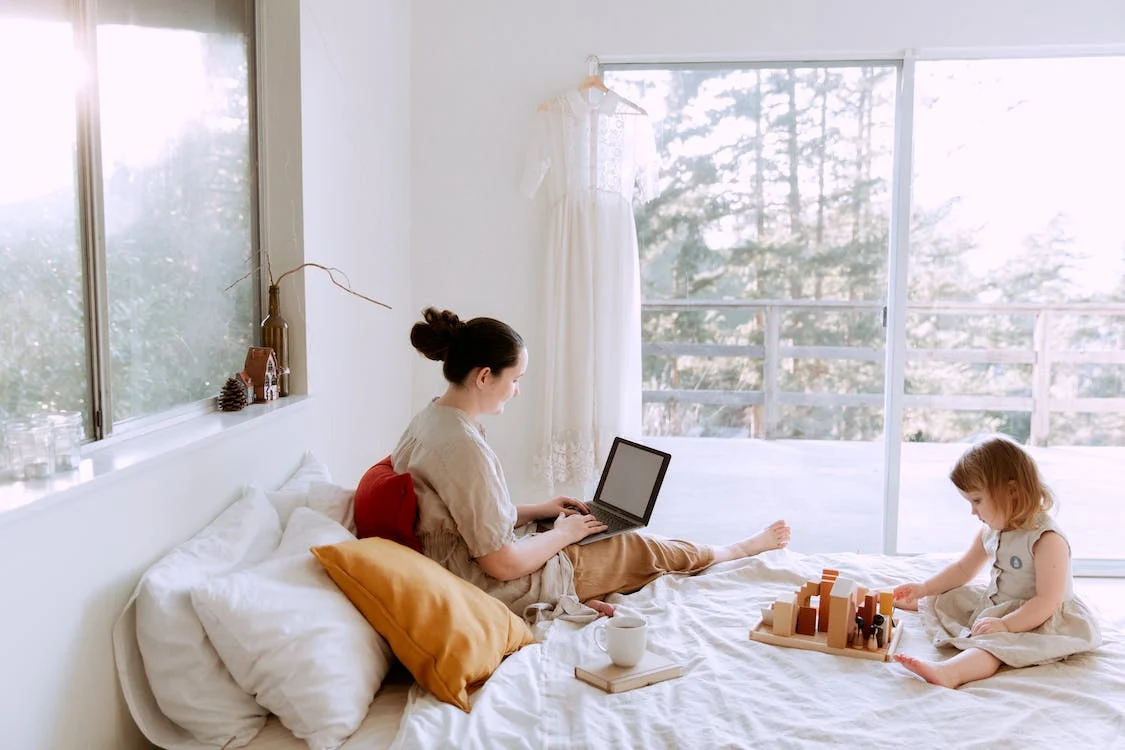girl playing with wooden blocks sitting on bed while mother using laptop