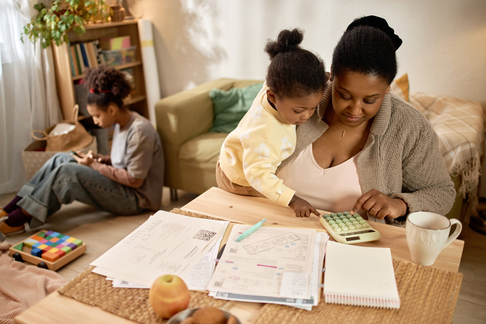 African American mother calculating household bills while holding child, teenager sitting nearby at home. Ideal for poverty, financial stress, budgeting, social support campaigns