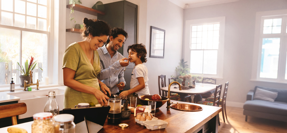 Playful dad feeding his son a slice of bread while his wife prepares breakfast. Family of three having fun together in the kitchen. Mom and dad are spending quality time with their son