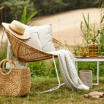 Stylish composition of outdoor garden on the lake with design rattan armchair, coffee table, plaid, pillows, drinks and elegant accessories. Summer chillout mood.